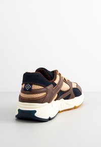 Brown, beige, and navy sneaker with textured fabric and leather panels, thick white sole, and a small "G" logo patch on the heel.