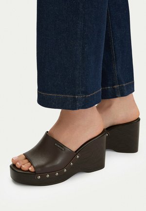 Pies con sandalias de cuña plataforma de color marrón oscuro con tachuelas, combinadas con pantalones vaqueros cortos azul oscuro.