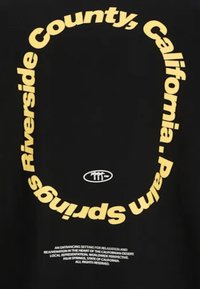 Camiseta negra con texto curvado en amarillo que dice "Riverside County, California" y "Palm Springs", con un pequeño logo en el centro.