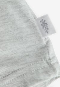 Ljust grått tygplagg med hörn och sömdetaljer samt en vit etikett med en diskret blommönstring. Mjuk textur, avslappnad design.