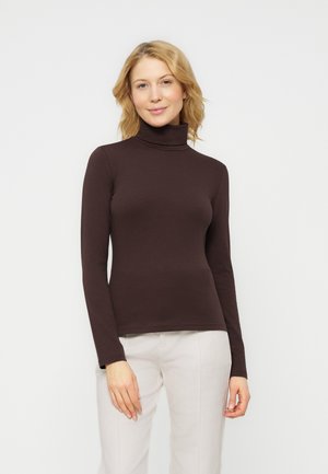 Femme blonde portant un col roulé marron foncé à manches longues et un pantalon beige clair, debout avec un léger sourire devant un fond uni.