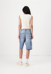 Mujer con cabello largo y oscuro que lleva puesto un top de punto blanco sin mangas, shorts de mezclilla azul claros hasta la rodilla, calcetas blancas y zapatillas blancas.