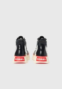 Zapatillas altas de charol negro con el logo rojo de SOREL en la parte posterior y suelas blancas, que presentan un exterior texturizado y un sistema de cordones.