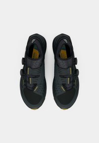 Czarne buty sportowe z teksturowaną cholewką, wyposażone w dwa regulowane pokrętła, żółte akcenty oraz gumową podeszwę. Smukły design zapewniający komfort.