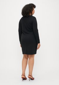 Μαύρο φόρεμα μακρυμάνικο με εφαρμοστή σιλουέτα, διαθέτοντας άνοιγμα στο πίσω μέρος και απαλό υφή. Συνδυασμένο με κόκκινα τακούνια.