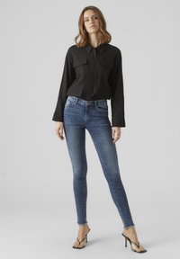 Černá zkrácená košile na knoflíky s dvěma náprsními kapsami, spárovaná s modrými skinny džínami s vysokým pasem a průhlednými sandály na vysokém podpatku.
