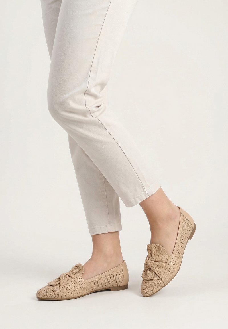 Beige flache Schuhe mit Perforationen und großer Schleifendetail, getragen zu hellbeigen, schmal geschnittenen, verkürzten Hosen vor schlichter Hintergrund.