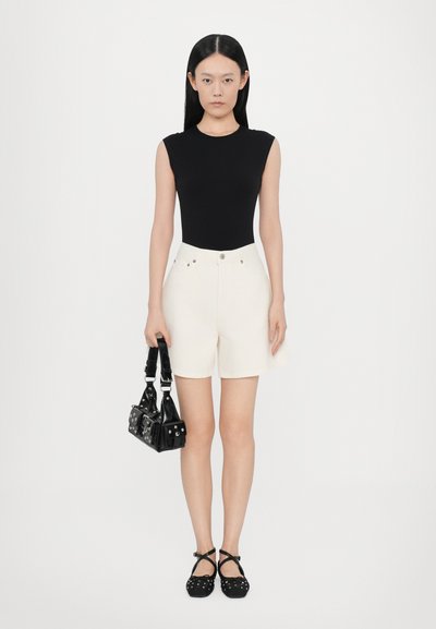 Μαύρο κορμάκι χωρίς μανίκια, κρεμ σορτς, μαύρη τσάντα με τρουκς, μαύρα παπούτσια με λουριά που σταυρώνονται· το μοντέλο στέκεται μπροστά σε λευκό φόντο.