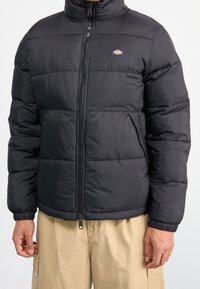 Uomo che indossa una giacca trapuntata nera con zip frontale e pantaloni beige, in piedi contro uno sfondo bianco semplice.