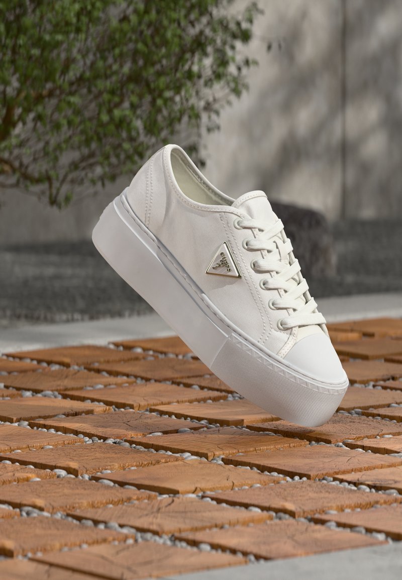 Baskets en toile blanche à semelle épaisse avec un logo triangulaire en métal, présentées en suspension au-dessus d'un sol en planches de bois et de petits graviers.