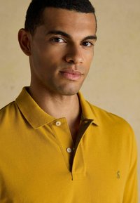 Sinine kollane polo särk, millel on krae, kaks nuppu, lühikesed varrukad ja rindkere kohal väike tikand.