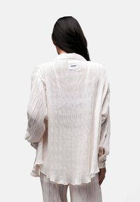 Camicetta leggera color crema con pieghe verticali, maniche lunghe e colletto. Presenta un'etichetta discreta sul retro e un design fluido.