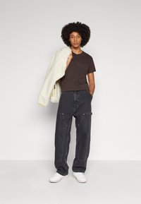 Brun, kortermet t-skjorte i kombinasjon med løse, mørkegrå denim cargo-bukser med frontlommer og hvite sneakers. Modell som holder en hvit jakke.