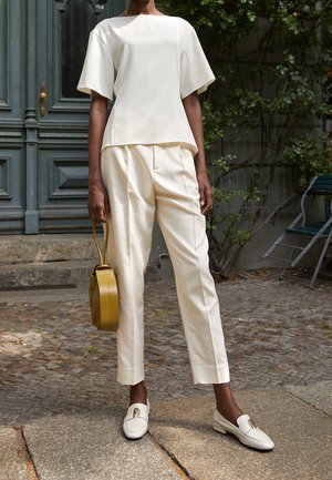 Roomkleurige, geplooide top gecombineerd met aansluitende, lichtbeige broek. Model draagt een ronde, olijfgroene handtas; draagt witte loafers met accenten.