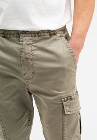 Olivgrüne Cargo-Hosen mit elastischem Bund, Seitentaschen und einer aufgesetzten Tasche am Oberschenkel. Der Stoff scheint glatt und leicht zu sein.