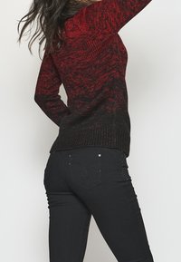 Femme aux longs cheveux foncés portant un pull rouge et noir à motifs et un jean noir ajusté, posant avec un bras levé.
