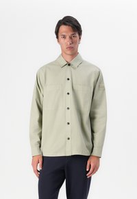 Lichtgroen button-up shirt met twee zijzakken, zwarte knopen en een patch op de linkermouw. Lange mouwen en een losse pasvorm.