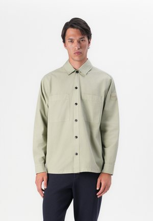 Light green button-up shirt with two front pockets, black buttons, and a patch on the left sleeve. Long sleeves and relaxed fit.
