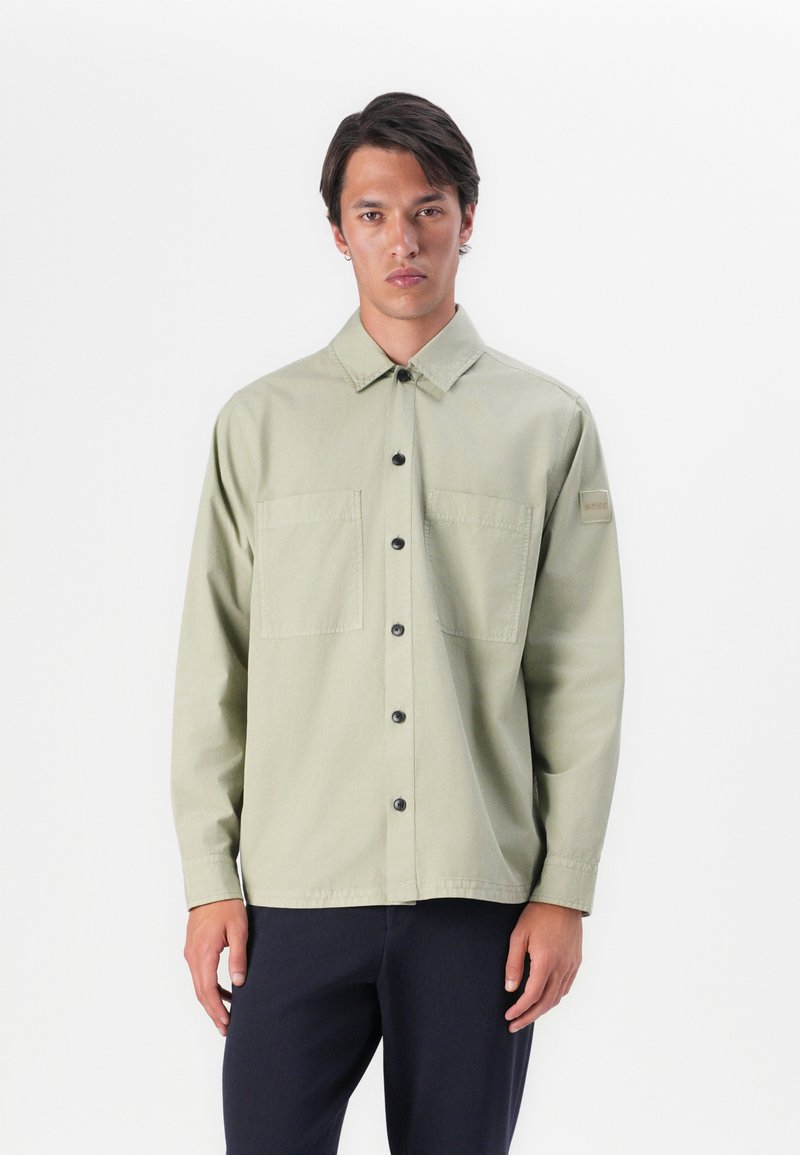 Lichtgroen button-up shirt met twee zijzakken, zwarte knopen en een patch op de linkermouw. Lange mouwen en een losse pasvorm.