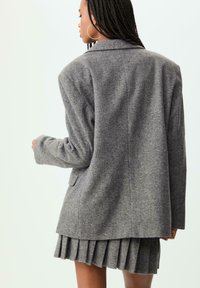 Blazer grigio in misto lana con colletto a rever, vestibilità su misura e due tasche, abbinato a una gonna plissettata grigia che mette in mostra un tessuto strutturato.