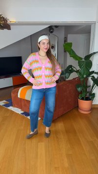 Γυναίκα με μακριά μαλλιά που φοράει ζακέτα με ζιγκ-ζαγκ σε ροζ, πορτοκαλί και πράσινο χρώμα και τζιν παντελόνι στέκεται στο σαλόνι κοντά σε καφέ καναπέ και ψηλό φυτεμένο φυτό.
