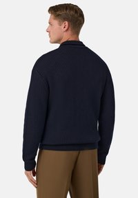 Mörkblå stickad cardigan med ribbad textur, med krage och långa ärmar, tillsammans med ljusbruna byxor.