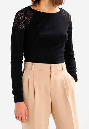 Mujer con un top negro de manga larga con detalles de encaje en los hombros y pantalones plisados beige de talle alto con las manos en los bolsillos.