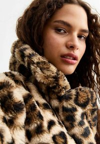 Cappotto in pelliccia sintetica con stampa leopardata, colletto alto e texture morbida; presenta macchie marroni scure su uno sfondo beige, evidenziando il materiale morbido.