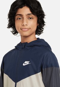 Jachetă de vânt bleu marin și crem cu glugă. Prezintă logo-ul Nike în alb pe piept. Textură netedă, închidere cu fermoar și panouri de culoare contrastantă.