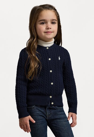 Ναυτικό κασκόλ με πλέξη καλωδίων και στρογγυλή λαιμόκοψη, με κουμπιά μπροστά και μικρό λογότυπο στο στήθος; συνδυασμένο με λευκό turtleneck από κάτω.