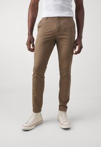 Pantalons slim-fit marron en mélange de coton, avec une taille plate, des poches standard, portés avec des baskets montantes blanches.