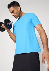 Blaues kurzärmeliges Sportshirt mit strukturierter Oberfläche, ausgestattet mit einem kleinen Logo. Getragen mit schwarzen Shorts und eine Hantel haltend.