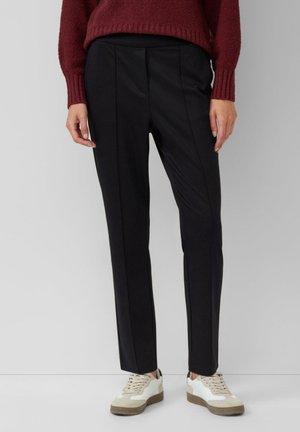 Personne portant un pantalon noir sur mesure, des baskets blanches et un pull en laine bordeaux, debout devant un fond gris clair uni.