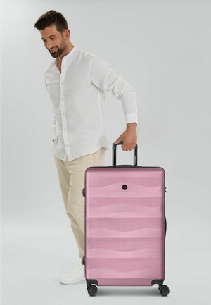 Mann in weißem Hemd und beigen Hosen hält den Griff eines großen pinkfarbenen Koffers mit vier Rollen vor grauem Hintergrund.