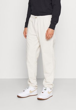 Mężczyzna ubrany w luźne białe sztruksowe spodnie, białe trampki z beżowym znakiem oraz czarną koszulę z długim rękawem, stojący na jednolitym tle.