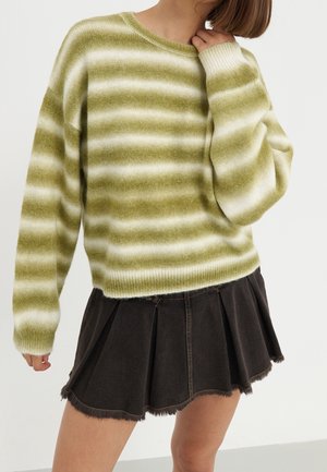 Persoon die een groen-wit gestreepte, pluizige trui draagt en een zwarte geplooide minirok met rafelige zoom tegen een effen achtergrond.