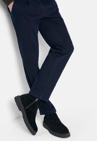 Schwarze Wildleder-Stiefeletten mit einem Reißverschluss, kombiniert mit dunkelblauen, geraden Hosen. Die Stoffe wirken weich mit einer sauberen Verarbeitung.