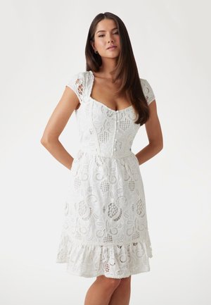Robe en dentelle blanche avec de courtes manches à capuchon, décolleté en scoop, corsage ajusté, détails de boutons et ourlet à volants présentant des motifs floraux.