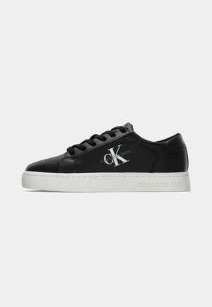 Czarne niskie trampki z białą podeszwą, czarnymi sznurowadłami i dużym logo "CK Calvin Klein Jeans" z boku i na podeszwie.