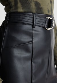 Jupe en cuir noir avec une taille côtelée, une boucle en or et une poche sur le côté. La texture semble lisse avec des plis doux.