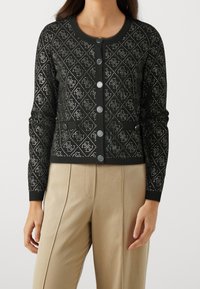 Cardigan noir avec un motif en dentelle, comportant des boutons en argent, un col rond, des manches longues et deux poches avant.