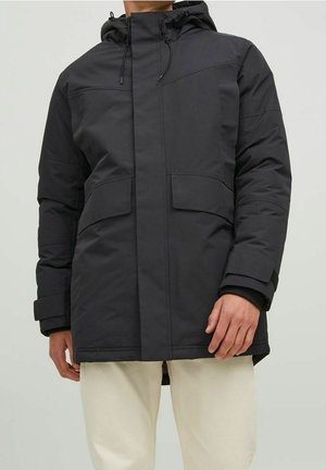 Mann trägt schwarze Winterjacke mit Kapuze und großen Fronttaschen sowie beige Hosen, steht vor einfarbigem Hintergrund.