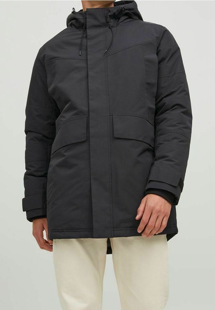 Homme portant une veste d'hiver noire à capuche avec de grandes poches avant et un pantalon beige, debout devant un fond uni.