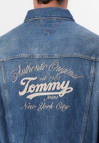 Denimjakke i mellomblå med brodert tekst: "Authentic Original Tommy Jeans est 1985 New York City" på baksiden. Klassisk design.
