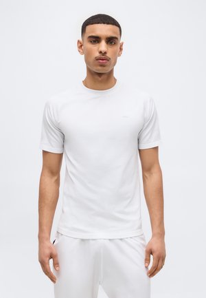 Fiatal férfi rövid hajú, egyszerű fehér rövid ujjú póló és fehér nadrág visel, világos háttér előtt áll.