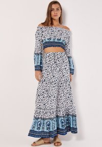 Top court décolleté avec un imprimé floral, associé à une jupe longue avec des volants et un ourlet à motifs bleus. Des sandales plates complètent le look.