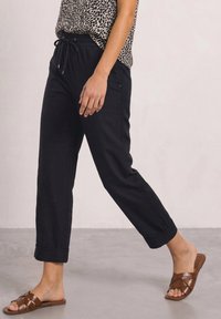 Person walking wearing black cuffed pants, animal print top, and brown woven leather slip-on sandals on gray floor.
