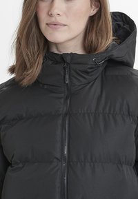 Chaqueta de plumas negra con cuello alto, cremallera frontal completa y capucha extraíble. La tela texturizada presenta secciones acolchadas horizontales.