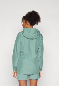 Giacca impermeabile verde con cappuccio, caratterizzata da una texture liscia, polsini elasticizzati e un fit casual, abbinata a pantaloni shorts coordinati.