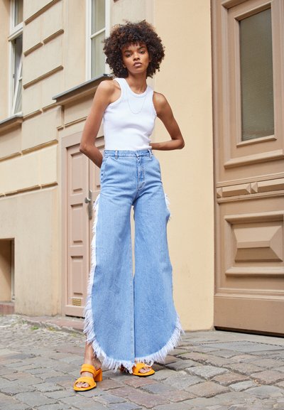 Himmelblaue weite Jeans mit fransigen Kanten, kombiniert mit einem weißen Tank-Top und orangefarbenen hohen Sandalen, vor einem Steinpflaster-Hintergrund.